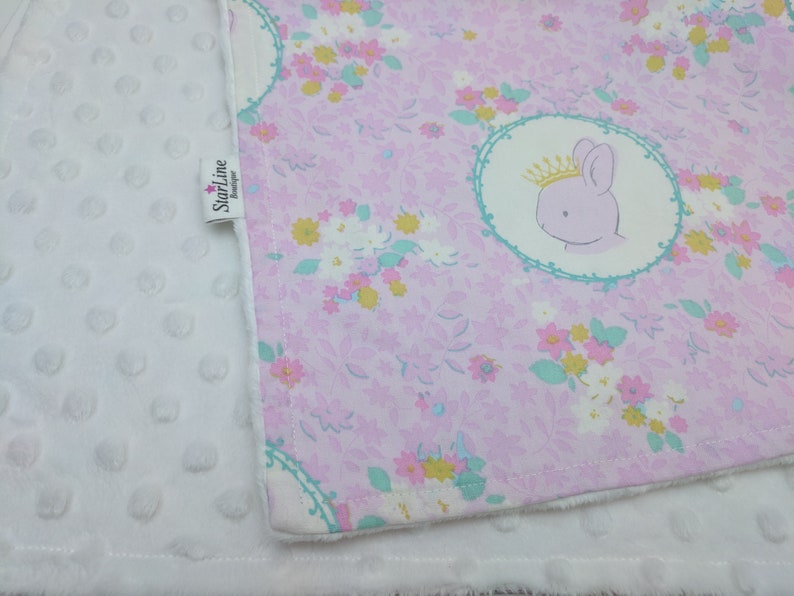 Puede incluir: Una manta de beb&eacute; blanca con un estampado floral rosa en un lado. El estampado floral presenta una corona y un conejito. La manta tiene una etiqueta que dice "Starline Designer".