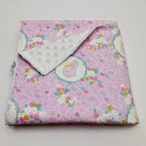 Puede incluir: Una manta de beb&eacute; rosa y blanca con un patr&oacute;n floral y un respaldo de minky blanco. La manta presenta un peque&ntilde;o conejito bordado con una corona.