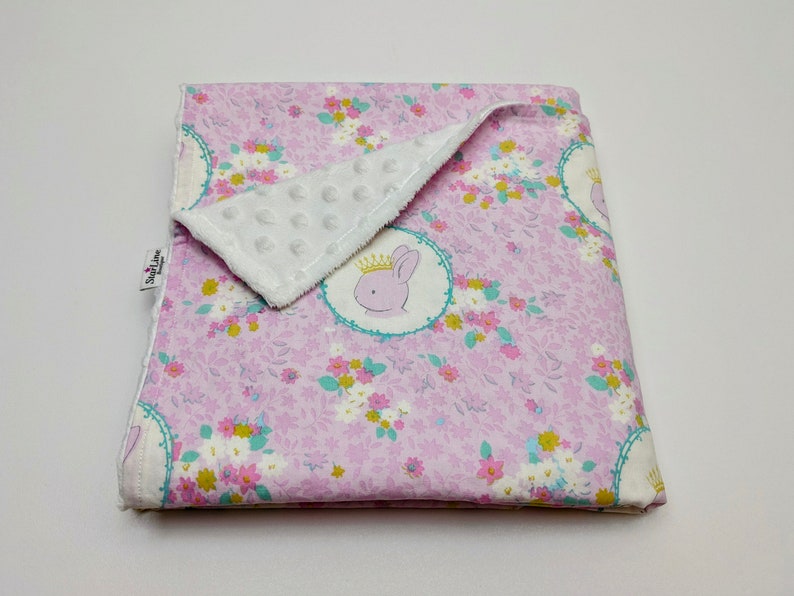 Puede incluir: Una manta de beb&eacute; rosa y blanca con un patr&oacute;n floral y un respaldo de minky blanco. La manta presenta un peque&ntilde;o conejito bordado con una corona.