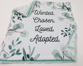 Puede incluir: Una manta para bebé blanca y verde con una textura suave y peluda en un lado. La manta presenta un diseño floral de estilo acuarela y el texto "Wanted. Chosen. Loved. Adopted."