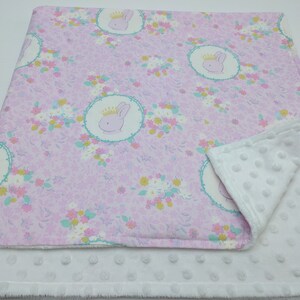 Puede incluir: Una manta de beb&eacute; rosa y blanca con un patr&oacute;n floral y una imagen repetida de un conejito con una corona. La manta est&aacute; hecha de dos capas de tela, una con una textura suave y mullida y la otra con una textura lisa de algod&oacute;n.