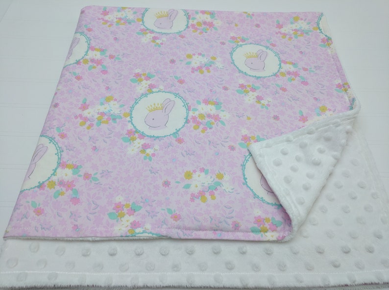 Puede incluir: Una manta de beb&eacute; rosa y blanca con un patr&oacute;n floral y una imagen repetida de un conejito con una corona. La manta est&aacute; hecha de dos capas de tela, una con una textura suave y mullida y la otra con una textura lisa de algod&oacute;n.