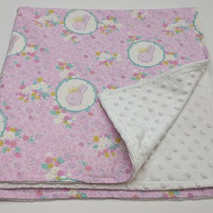 Puede incluir: Una manta de beb&eacute; rosa con un respaldo de minky blanco. La manta presenta un patr&oacute;n floral con un dise&ntilde;o repetido de un conejito con una corona.