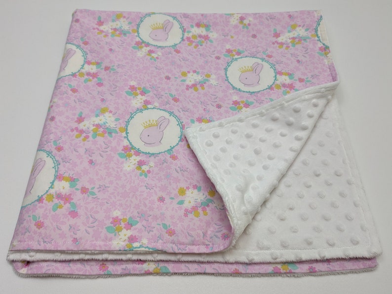 Puede incluir: Una manta de beb&eacute; rosa con un respaldo de minky blanco. La manta presenta un patr&oacute;n floral con un dise&ntilde;o repetido de un conejito con una corona.
