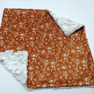 May include: A soft, plush blanket with a white floral pattern on a rust-colored background. The blanket is reversible with a white faux fur side.