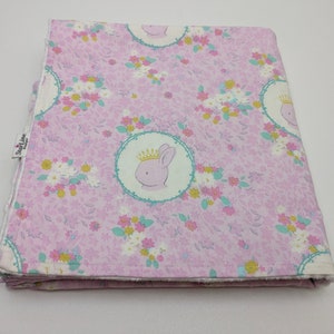 Puede incluir: Una manta de beb&eacute; rosa con un patr&oacute;n floral y una imagen repetida de un conejito con una corona. La manta tiene un borde blanco y el texto "SweetLine Designs" est&aacute; impreso en el lateral.
