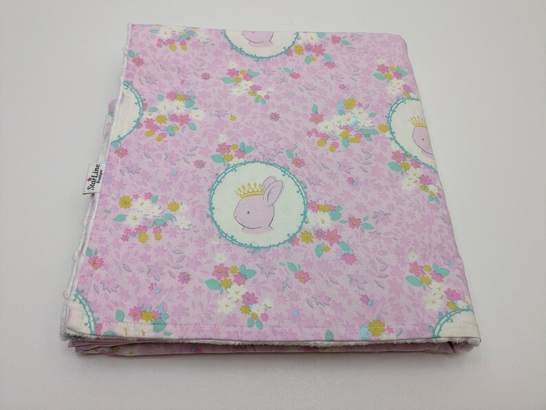 Puede incluir: Una manta de beb&eacute; rosa con un patr&oacute;n floral y una imagen repetida de un conejito con una corona. La manta tiene un borde blanco y el texto "SweetLine Designs" est&aacute; impreso en el lateral.