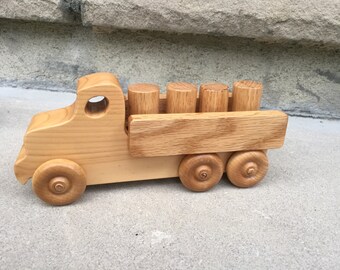 May include: A wooden toy truck with a flatbed and five logs in the back. The truck is made of light brown wood and has four wheels.