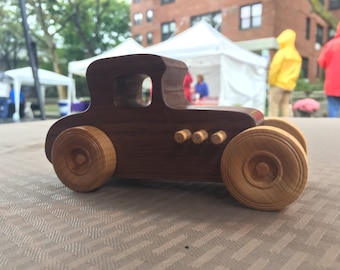 May include: A wooden toy car with brown body and light brown wheels. The car has a detailed design with a rounded hood and a grill with five small holes.