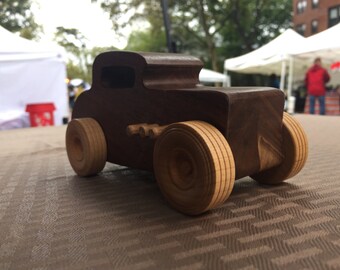 May include: A wooden toy car with brown paint and light brown wheels. The car is sitting on a brown patterned surface.