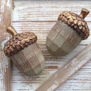 Two artificial acorns. The top of each acorn is a textured, metallic gold color, while the body is a light brown with a wood grain pattern. Fall Table Decor Acorn, Fall Mantel Decoration, Centerpiece Decor, Acorn Fall Home Decor, Thanksgiving Mantel Decor, Artificial Acorn Decor