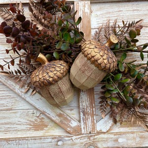 Two artificial acorns. The top of each acorn is a textured, metallic gold color, while the body is a light brown with a wood grain pattern. Fall Table Decor Acorn, Fall Mantel Decoration, Centerpiece Decor, Acorn Fall Home Decor, Thanksgiving Mantel Decor, Artificial Acorn Decor