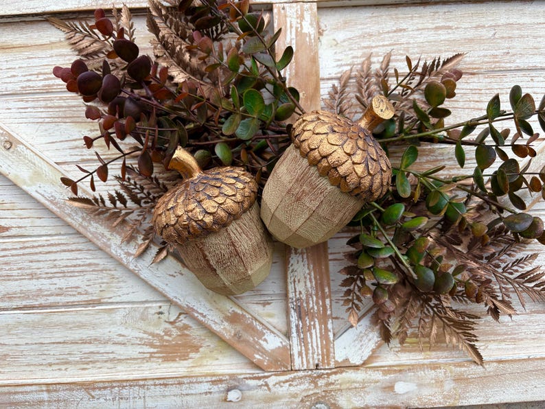 Two artificial acorns. The top of each acorn is a textured, metallic gold color, while the body is a light brown with a wood grain pattern. Fall Table Decor Acorn, Fall Mantel Decoration, Centerpiece Decor, Acorn Fall Home Decor, Thanksgiving Mantel Decor, Artificial Acorn Decor
