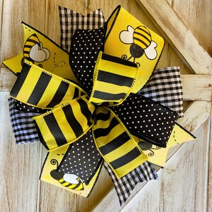 May include: A yellow and black bee-themed bow with black and white polka dot, gingham, and striped ribbon. The bow is tied in a large, decorative knot.