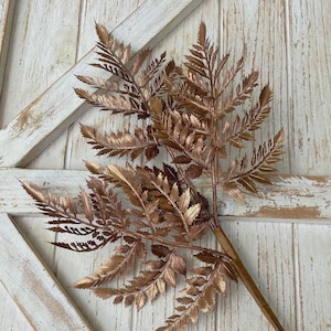 May include: A bronze-colored artificial fern branch with detailed, layered leaves. The faux foliage has a metallic finish and is set against a rustic, white-painted wooden background. This decorative piece is suitable for home decor and floral arrangements.