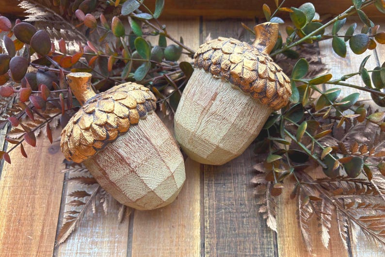 Two artificial acorns. The top of each acorn is a textured, metallic gold color, while the body is a light brown with a wood grain pattern. Fall Table Decor Acorn, Fall Mantel Decoration, Centerpiece Decor, Acorn Fall Home Decor, Thanksgiving Mantel Decor, Artificial Acorn Decor