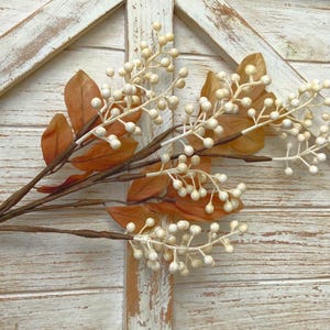 May include: A decorative floral stem featuring brown branches, orange leaves, and clusters of small, off-white berries. The artificial plant is set against a rustic, white-painted wooden background, evoking a fall or harvest theme.