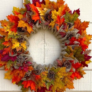 Op de afbeelding: Een herfstkrans met jute en kunstbladeren in tinten rood, oranje, geel en groen. De krans is versierd met kleine dennenappels.