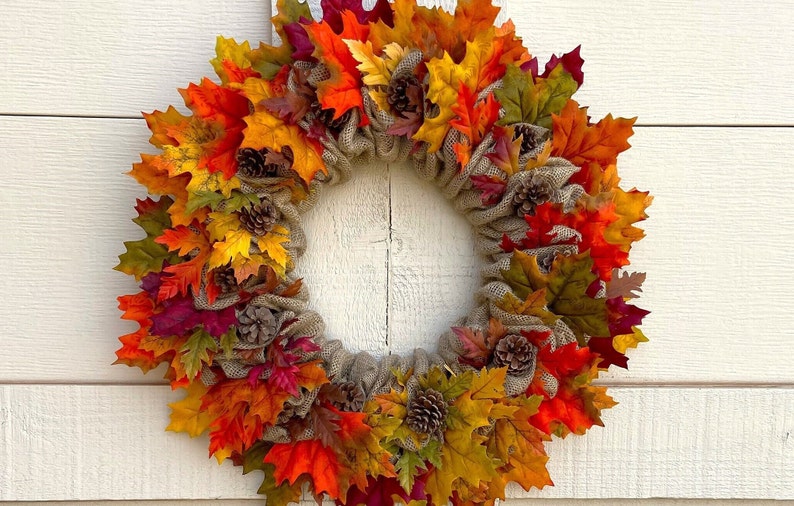 Op de afbeelding: Een herfstkrans met jute en kunstbladeren in tinten rood, oranje, geel en groen. De krans is versierd met kleine dennenappels.