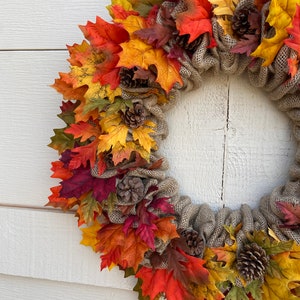 Op de afbeelding: Een jutekrans versierd met herfstbladeren in tinten rood, oranje, geel en bruin. De krans is versierd met kleine dennenappels.