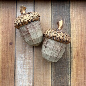 Two artificial acorns. The top of each acorn is a textured, metallic gold color, while the body is a light brown with a wood grain pattern. Fall Table Decor Acorn, Fall Mantel Decoration, Centerpiece Decor, Acorn Fall Home Decor, Thanksgiving Mantel Decor, Artificial Acorn Decor