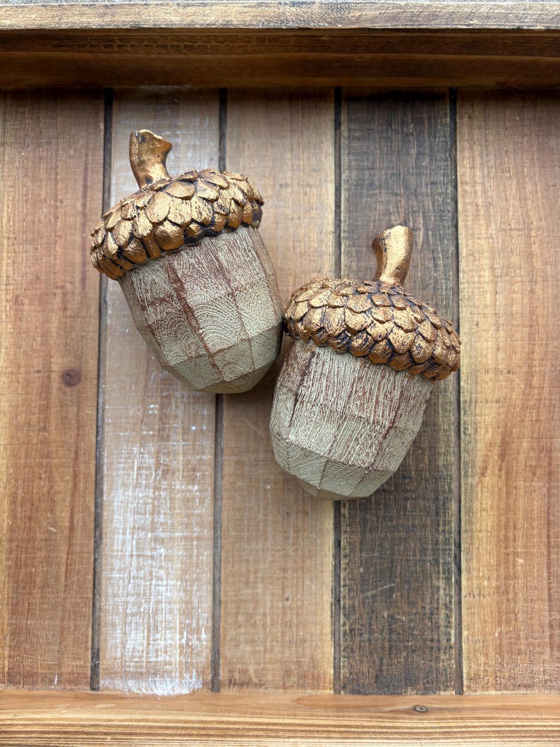 Two artificial acorns. The top of each acorn is a textured, metallic gold color, while the body is a light brown with a wood grain pattern. Fall Table Decor Acorn, Fall Mantel Decoration, Centerpiece Decor, Acorn Fall Home Decor, Thanksgiving Mantel Decor, Artificial Acorn Decor