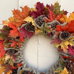 Op de afbeelding: Een herfstkrans met jute en kunstmatige esdoornbladeren in tinten rood, oranje, geel en bruin. De krans is versierd met dennenappels.