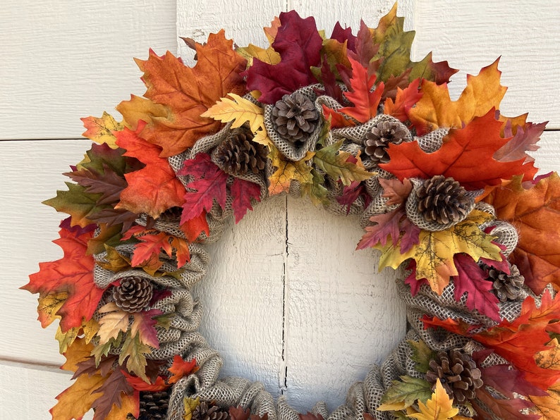 Op de afbeelding: Een herfstkrans met jute en kunstmatige esdoornbladeren in tinten rood, oranje, geel en bruin. De krans is versierd met dennenappels.