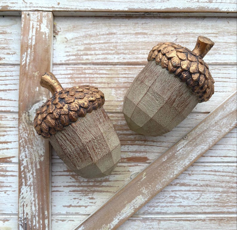 Two artificial acorns. The top of each acorn is a textured, metallic gold color, while the body is a light brown with a wood grain pattern. Fall Table Decor Acorn, Fall Mantel Decoration, Centerpiece Decor, Acorn Fall Home Decor, Thanksgiving Mantel Decor, Artificial Acorn Decor