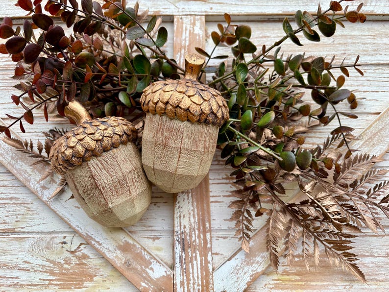 Two artificial acorns. The top of each acorn is a textured, metallic gold color, while the body is a light brown with a wood grain pattern. Fall Table Decor Acorn, Fall Mantel Decoration, Centerpiece Decor, Acorn Fall Home Decor, Thanksgiving Mantel Decor, Artificial Acorn Decor