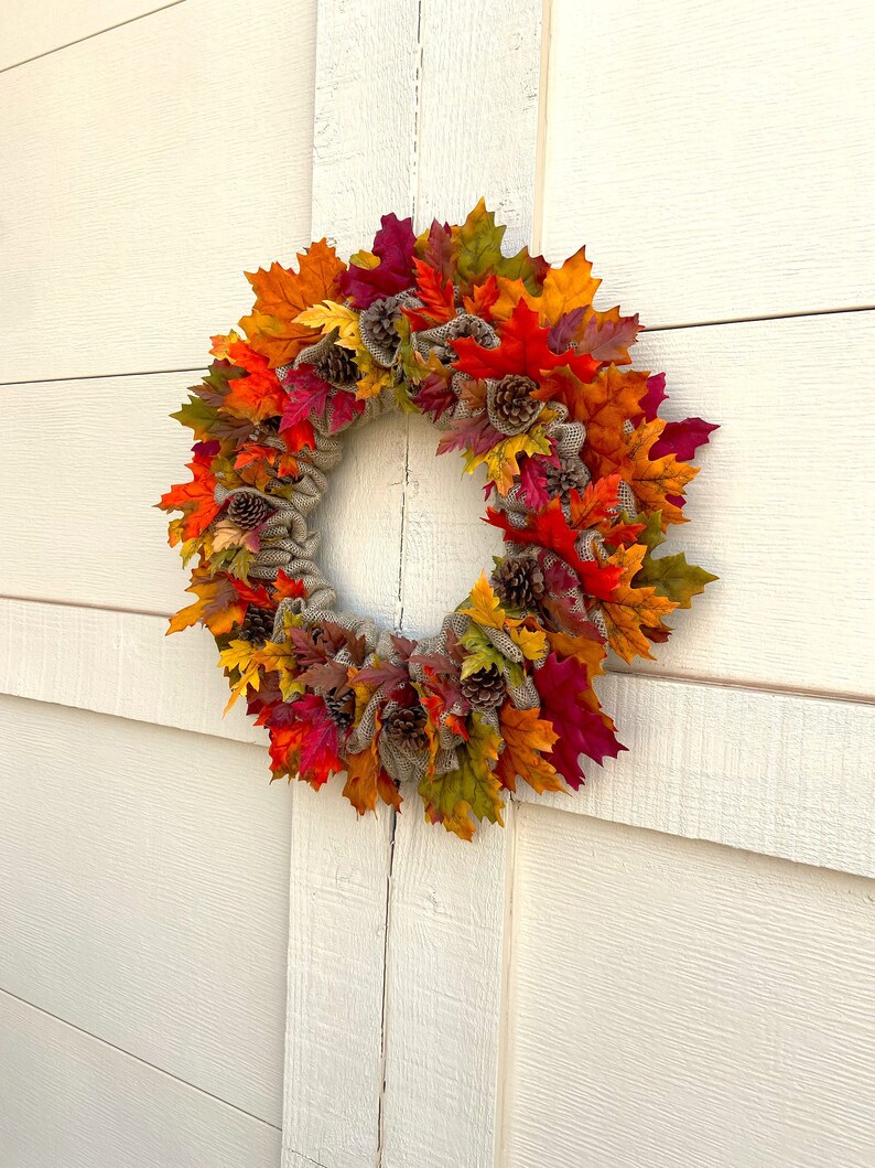Op de afbeelding: Een herfstkrans met jute en kunstmatige esdoornbladeren in tinten rood, oranje, geel en groen. De krans is versierd met dennenappels.