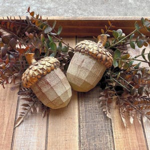 Two artificial acorns. The top of each acorn is a textured, metallic gold color, while the body is a light brown with a wood grain pattern. Fall Table Decor Acorn, Fall Mantel Decoration, Centerpiece Decor, Acorn Fall Home Decor, Thanksgiving Mantel Decor, Artificial Acorn Decor