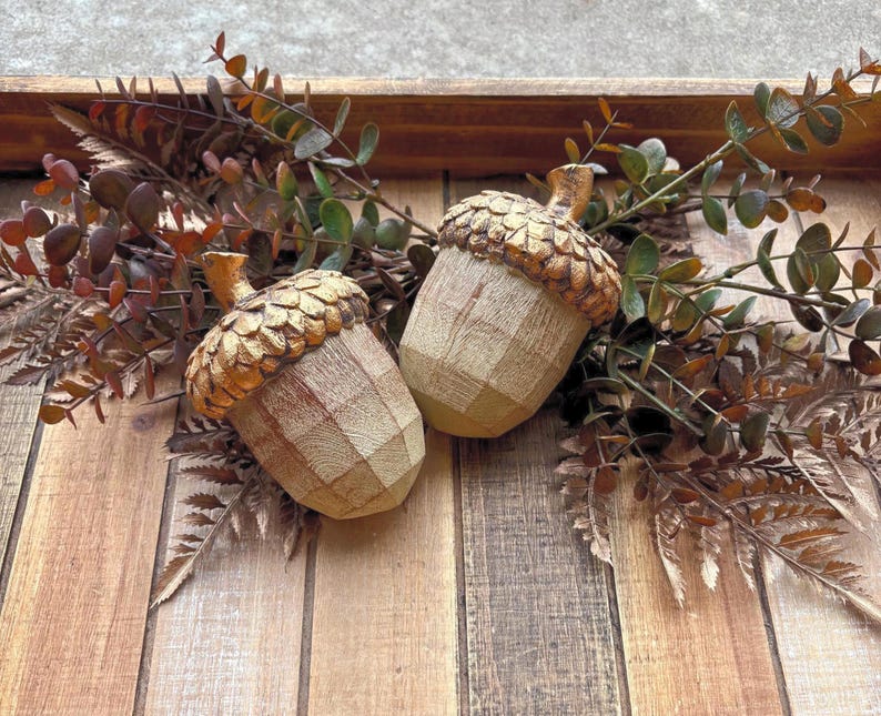 Two artificial acorns. The top of each acorn is a textured, metallic gold color, while the body is a light brown with a wood grain pattern. Fall Table Decor Acorn, Fall Mantel Decoration, Centerpiece Decor, Acorn Fall Home Decor, Thanksgiving Mantel Decor, Artificial Acorn Decor