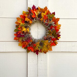 Op de afbeelding: Een herfstkrans met jute en kunstmatige bladeren in tinten rood, oranje, geel en groen. De krans is versierd met dennenappels.