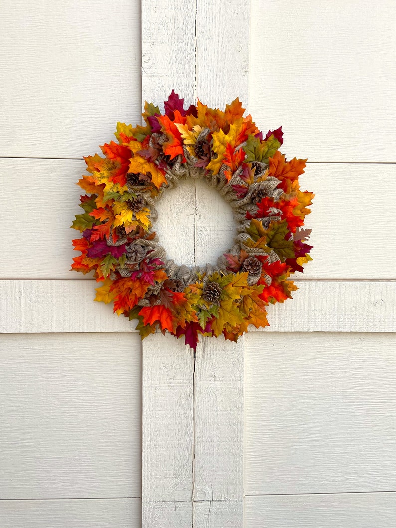 Op de afbeelding: Een herfstkrans met jute en kunstmatige bladeren in tinten rood, oranje, geel en groen. De krans is versierd met dennenappels.