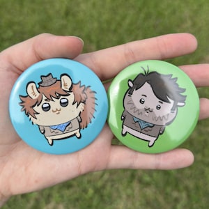 Puede incluir: Dos chapas circulares con ilustraciones de dibujos animados. La chapa izquierda es azul claro con una criatura de pelo castaño con sombrero y bufanda. La derecha es verde claro con una criatura de pelo oscuro con chaqueta marrón y bufanda.