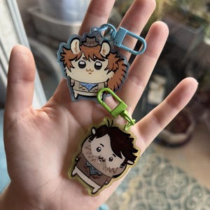 May include: Two acrylic keychains featuring cartoon character illustrations. The top keychain has a light blue clasp and depicts a brown-haired creature in a hat. The bottom keychain has a lime green clasp and shows a dark-haired character in a brown coat.