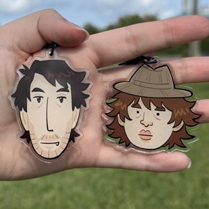 Puede incluir: Dos llaveros de acrílico transparente con cabezas de personajes ilustradas. El llavero de la izquierda muestra a una persona con cabello castaño oscuro y barba incipiente. El de la derecha muestra a una persona con sombrero fedora beige.