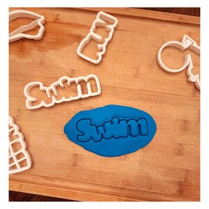 Op de afbeelding: Een blauw stuk modelleerklei met het woord "Swim" erin gedrukt. Er liggen verschillende witte plastic koekjesvormen op een houten oppervlak, waaronder een "Swim" koekjesvorm.