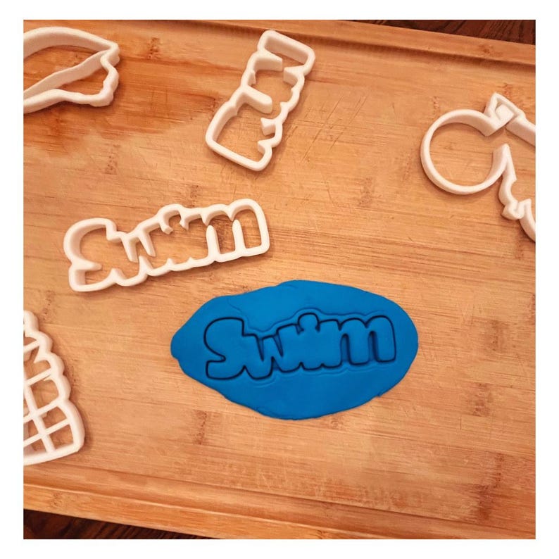 Op de afbeelding: Een blauw stuk modelleerklei met het woord "Swim" erin gedrukt. Er liggen verschillende witte plastic koekjesvormen op een houten oppervlak, waaronder een "Swim" koekjesvorm.