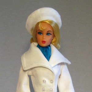 May include: A white doll wearing a white coat with a large collar, a white hat, and blue scarf. The doll is holding a white handbag and is standing on a clear plastic stand.