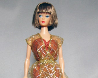 May include: A vintage Barbie doll wearing a red and gold dress with a sheer overlay. The dress has a geometric pattern and the doll has blue eyes and brown hair.