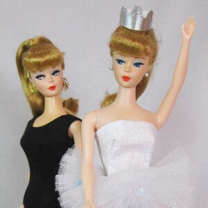 May include: Two Barbie dolls, one wearing a black leotard and the other wearing a white tulle tutu and a silver crown.