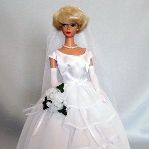May include: A white bridal gown with a satin bodice and a tulle skirt with multiple layers of ruffles. The bride is holding a bouquet of white flowers with green leaves.