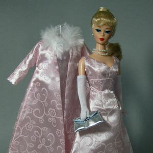 May include: A pink satin dress with a floral pattern and a matching pink satin coat with a white fur collar. The dress has a fitted bodice and a full skirt. The coat has a long, flowing silhouette. The doll is wearing white gloves and a silver clutch.