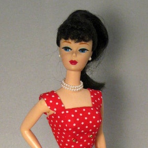 May include: A vintage doll wearing a red dress with white polka dots. The dress has a square neckline, gold buttons, and a fitted silhouette. The doll has black hair, blue eyes, and a pearl necklace. She is wearing red shoes.