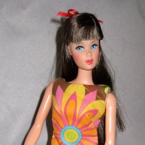 May include: A vintage doll with long brown hair, wearing a brown, pink, and yellow floral dress with a ruffled hem. The doll has blue eyes and is wearing orange shoes.
