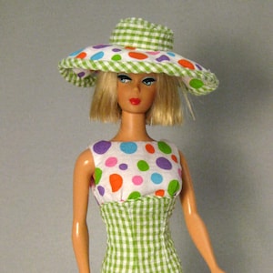 May include: A doll wearing a green and white gingham dress with a white background and colorful polka dots. The doll is wearing a green and white gingham hat.