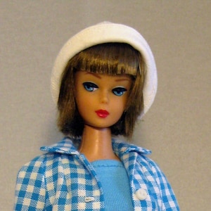 May include: A doll wearing a blue and white gingham jacket, blue pants, and a white hat. The doll has brown hair and blue eyes.