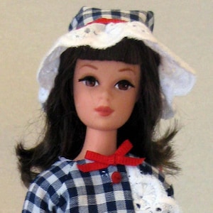 May include: A doll wearing a blue and white gingham dress with a white lace overlay, a red bow, and a white hat with a blue and white gingham trim. The doll is wearing red shoes and is standing on a clear display stand.